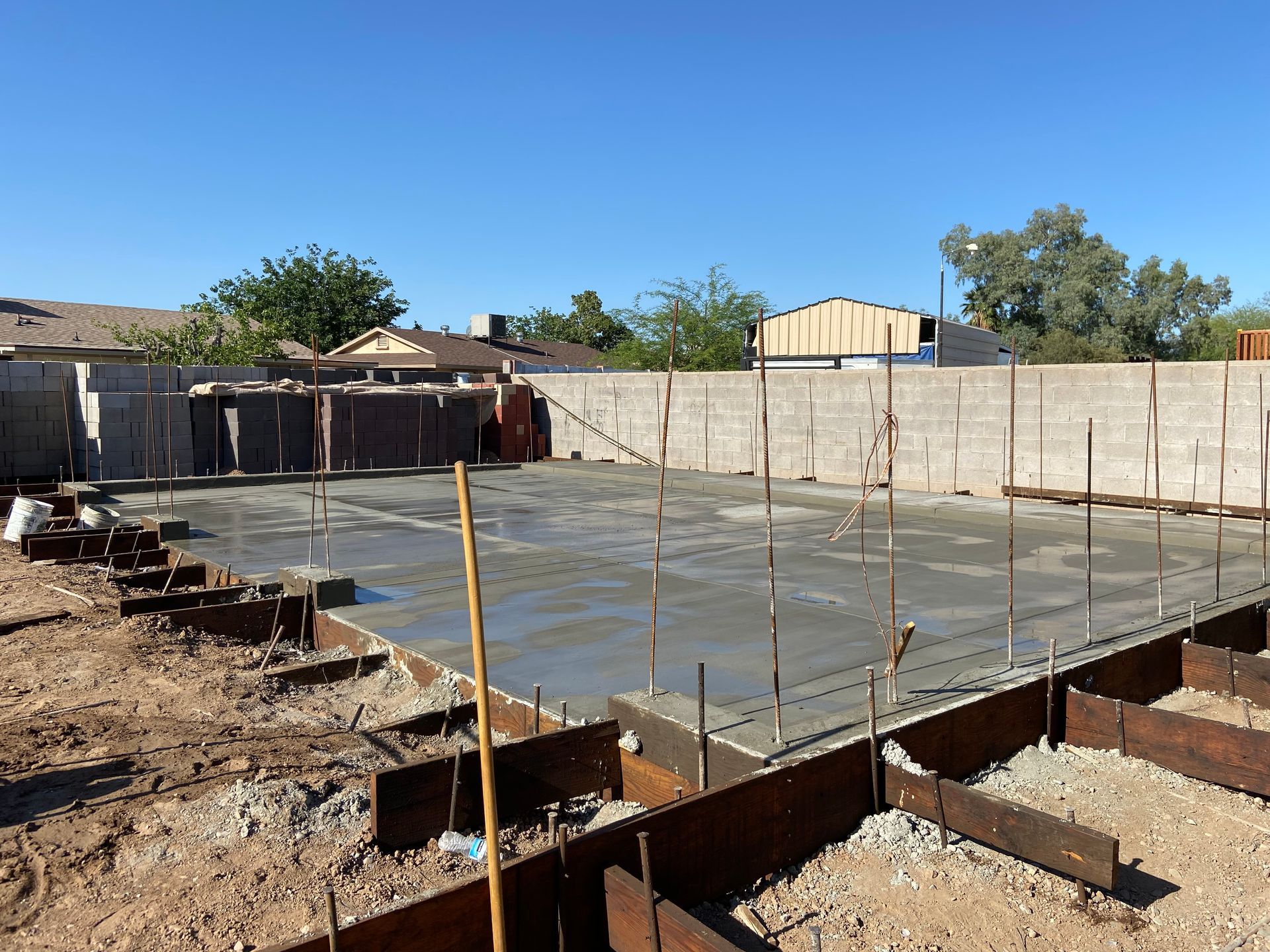 Concrete slab foundation under construction in a dirt lot, with rebar and wood forms beside a block wall.