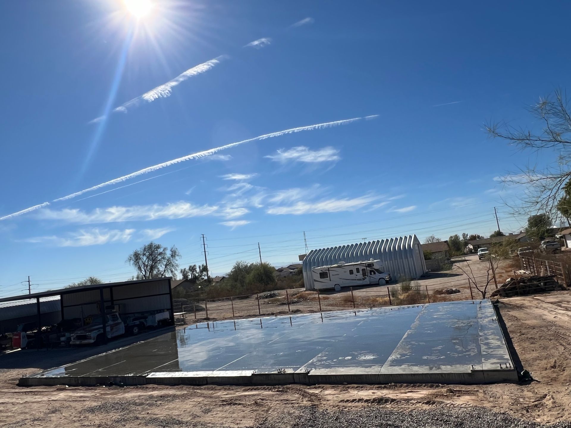 Desert yard with a reflective pool, solar panels, and bright blue sky with sun and contrails.