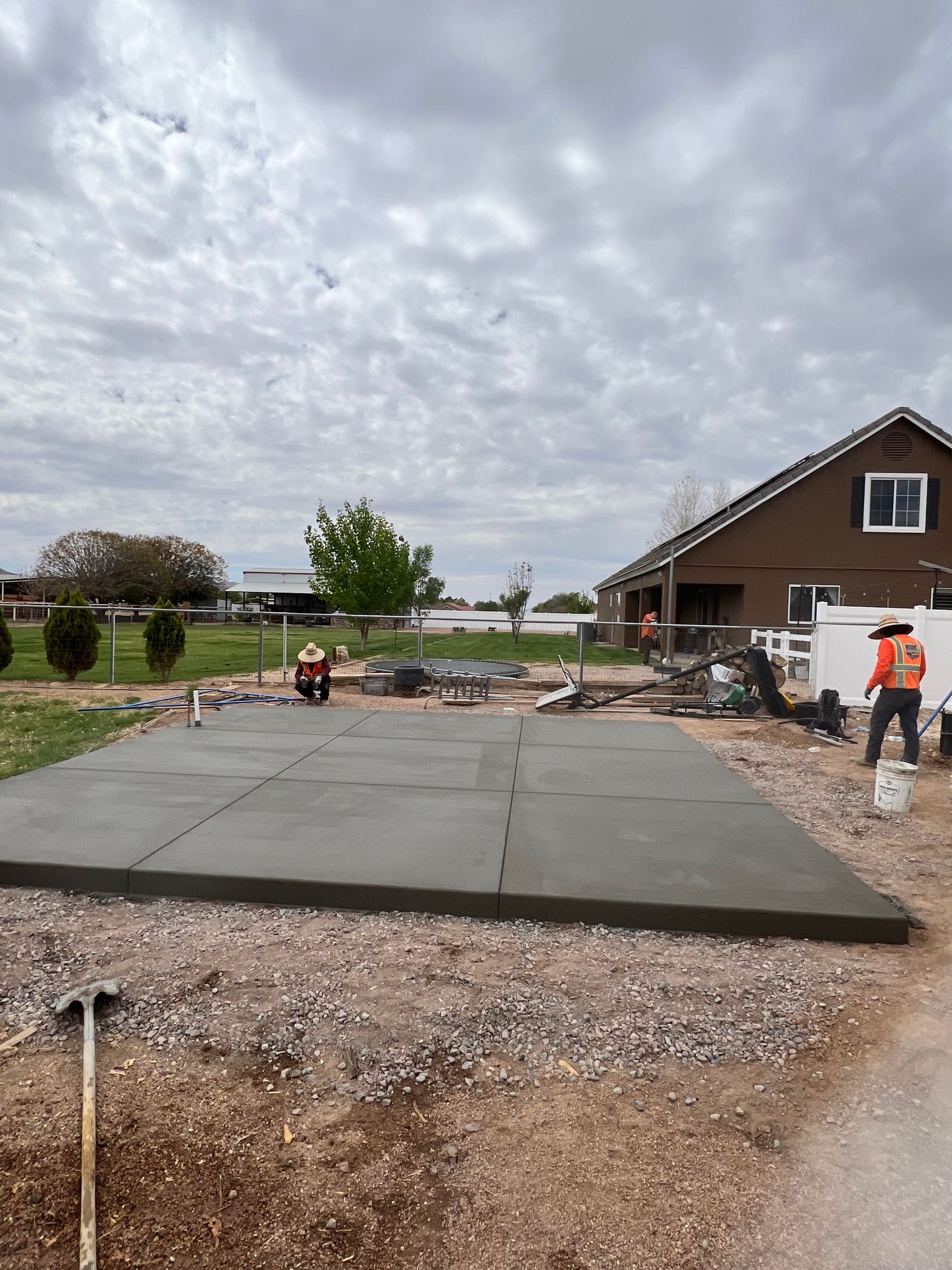 Workers pouring a concrete slab beside a brown house under a cloudy sky.