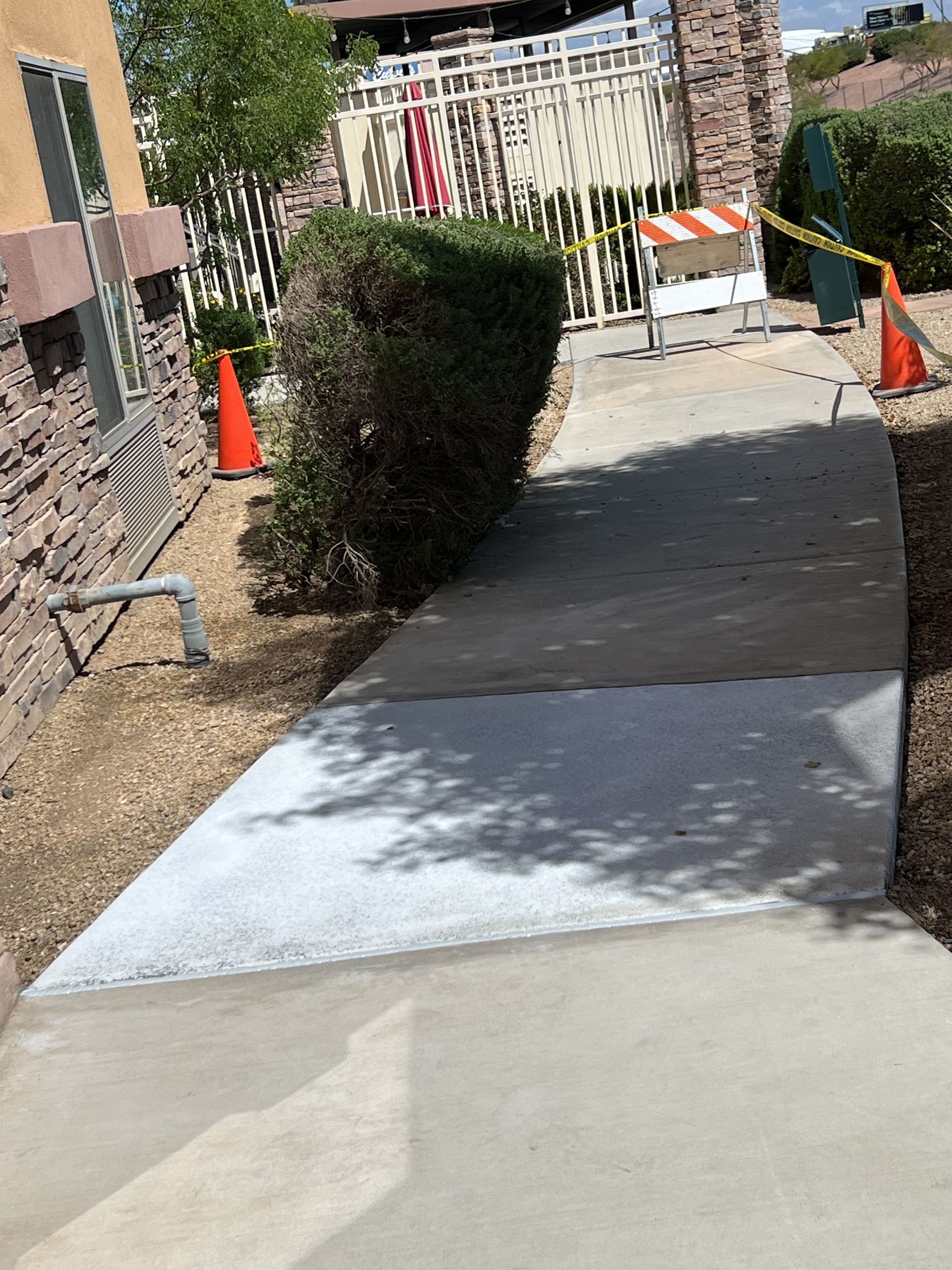 Narrow sidewalk under construction with orange cones, barriers, and a trimmed bush along a building side