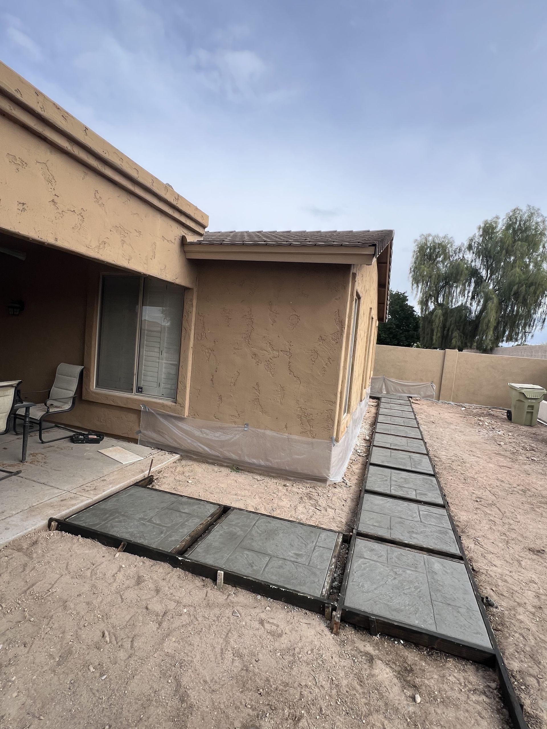 Concrete paver walkway beside a tan stucco house under construction, with gravel yard and blue sky.