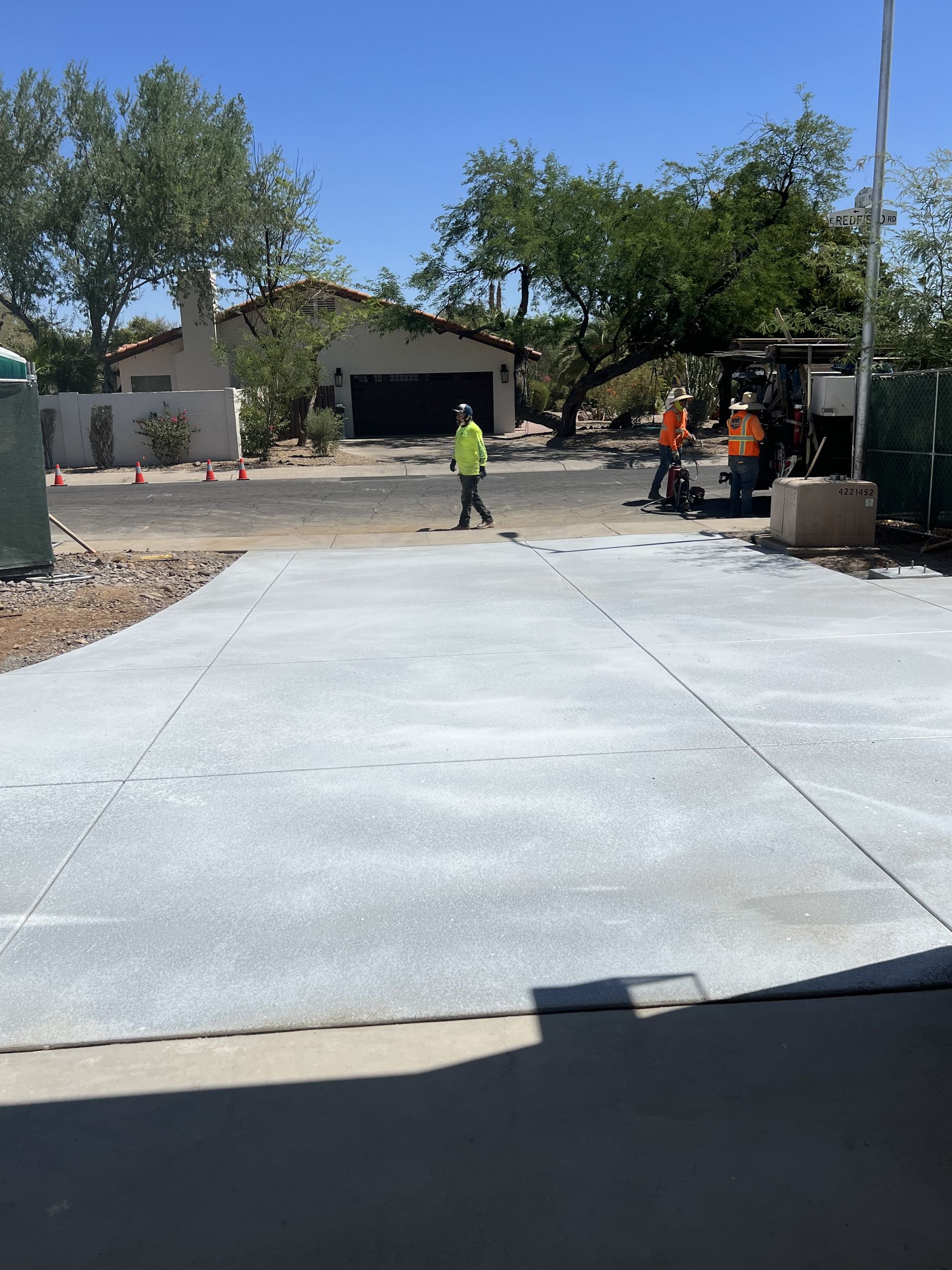 Concrete driveway under construction with workers, trees, and a house in the background