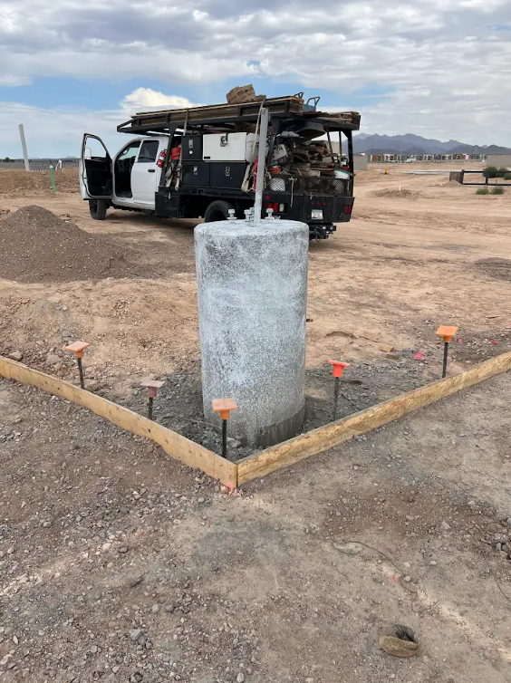 Concrete utility pedestal in a dirt lot beside an open work vehicle and wooden border stakes under a cloudy sky