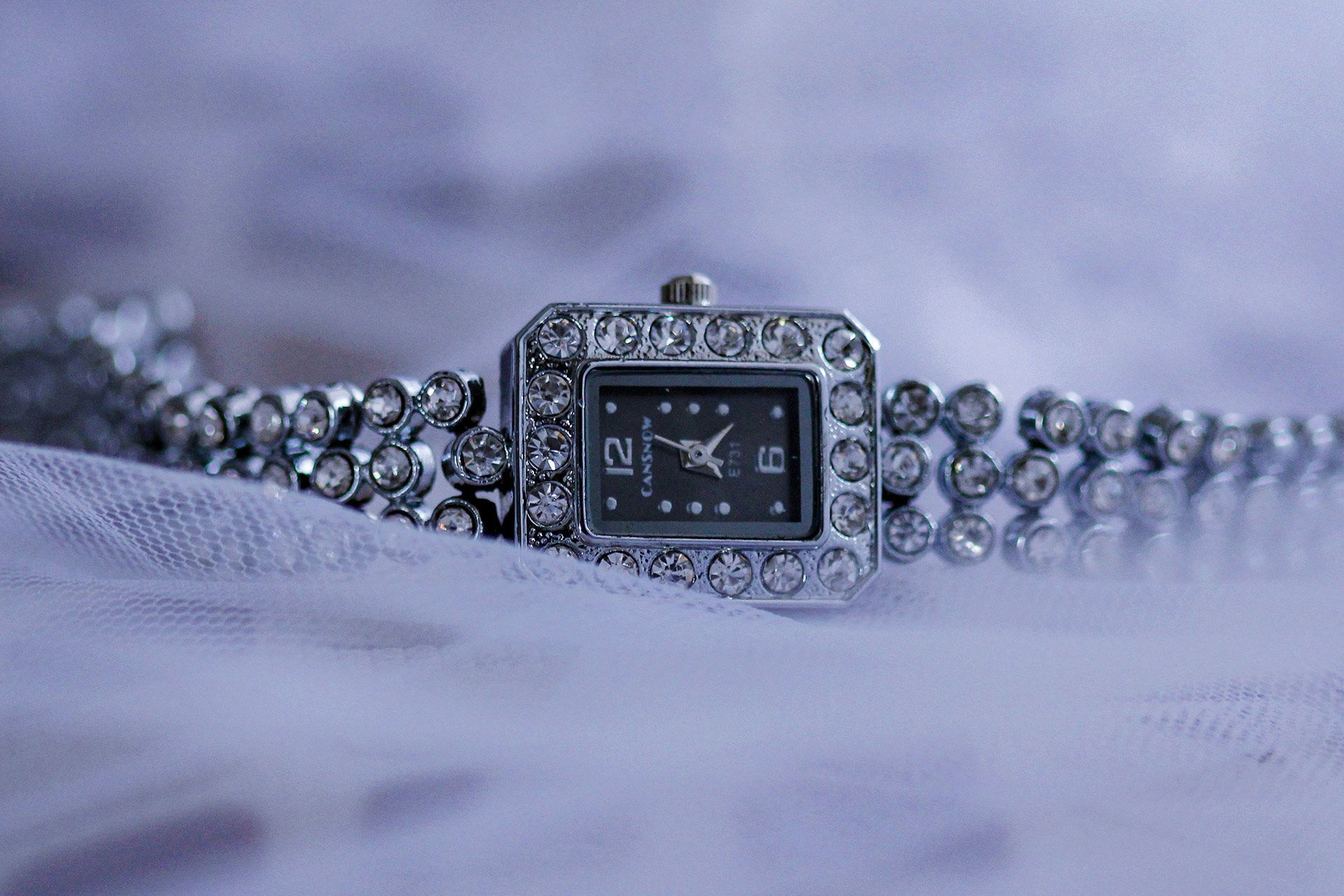 Silver jeweled wristwatch with square black face on a soft lavender fabric background