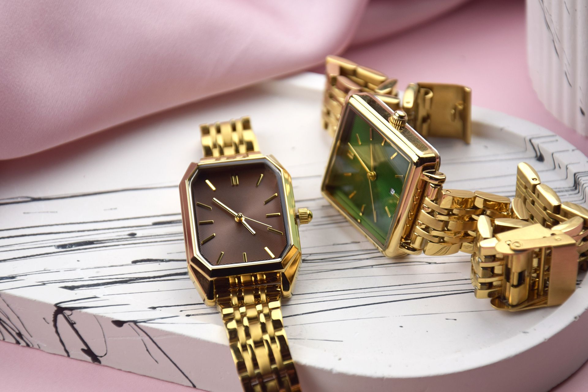 Two gold wristwatches on a white surface with pink fabric background