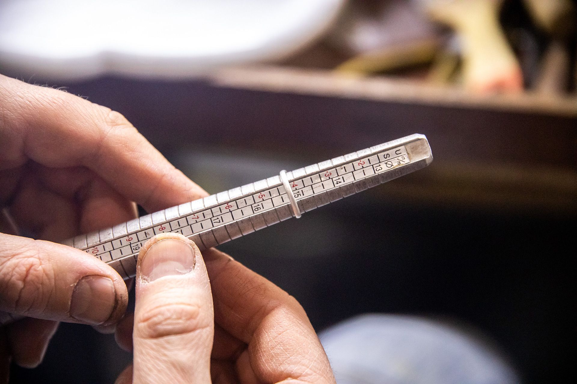 Hands holding a metal ring sizer with engraved measurements over a blurred work surface