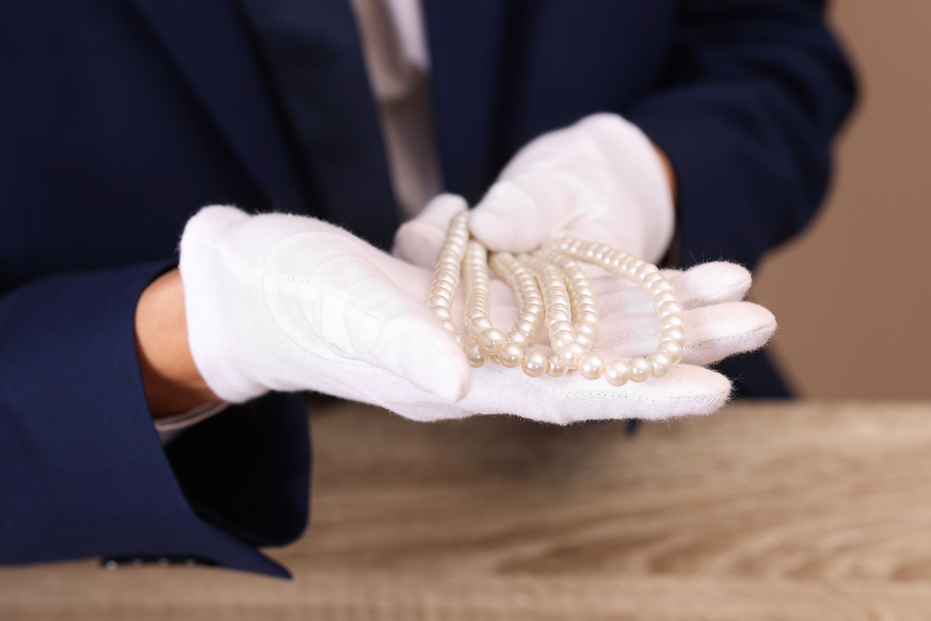 White-gloved hands holding a strand of pearls over a wooden table