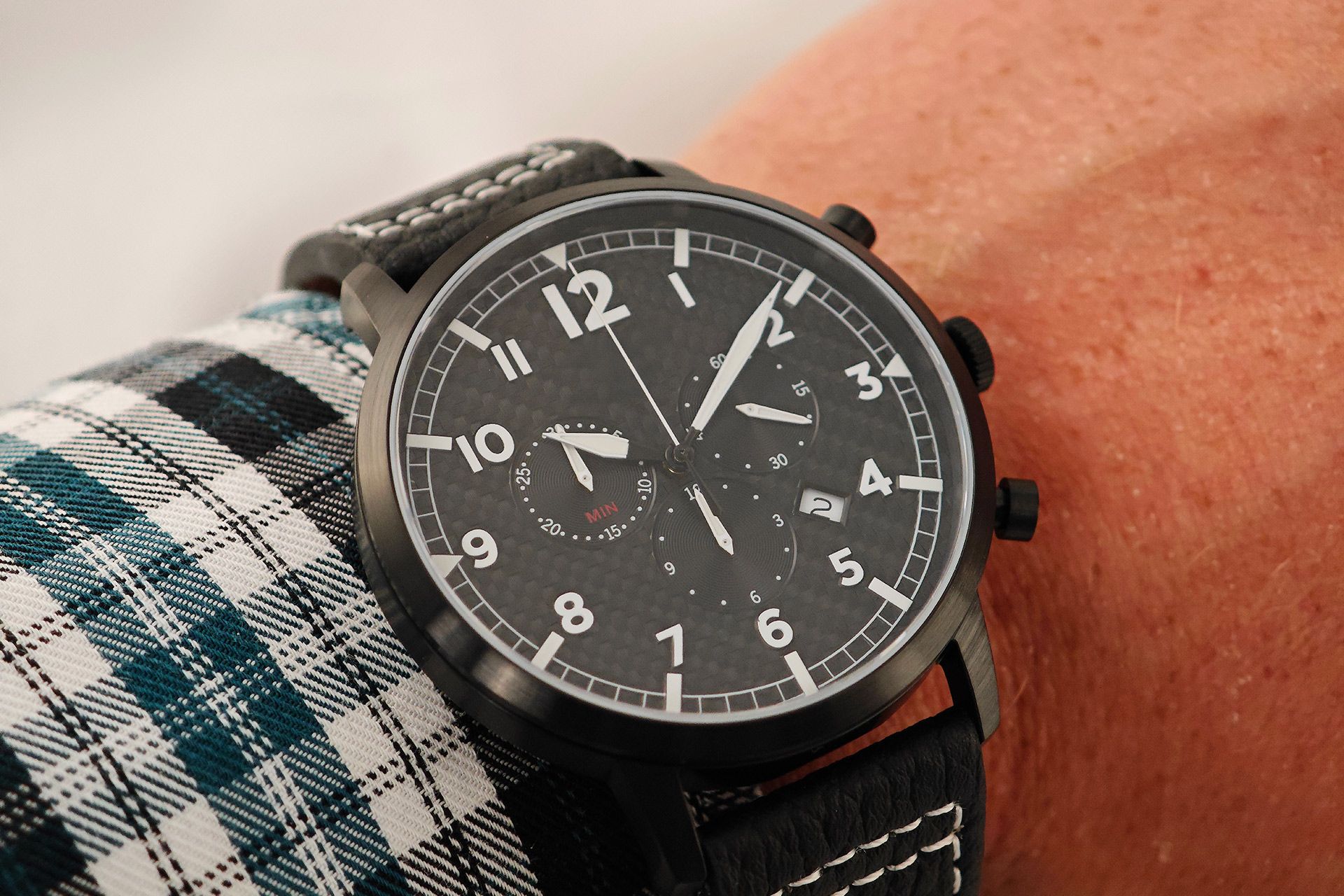 Black wristwatch with a dark dial on a person’s wrist, worn over a plaid shirt cuff