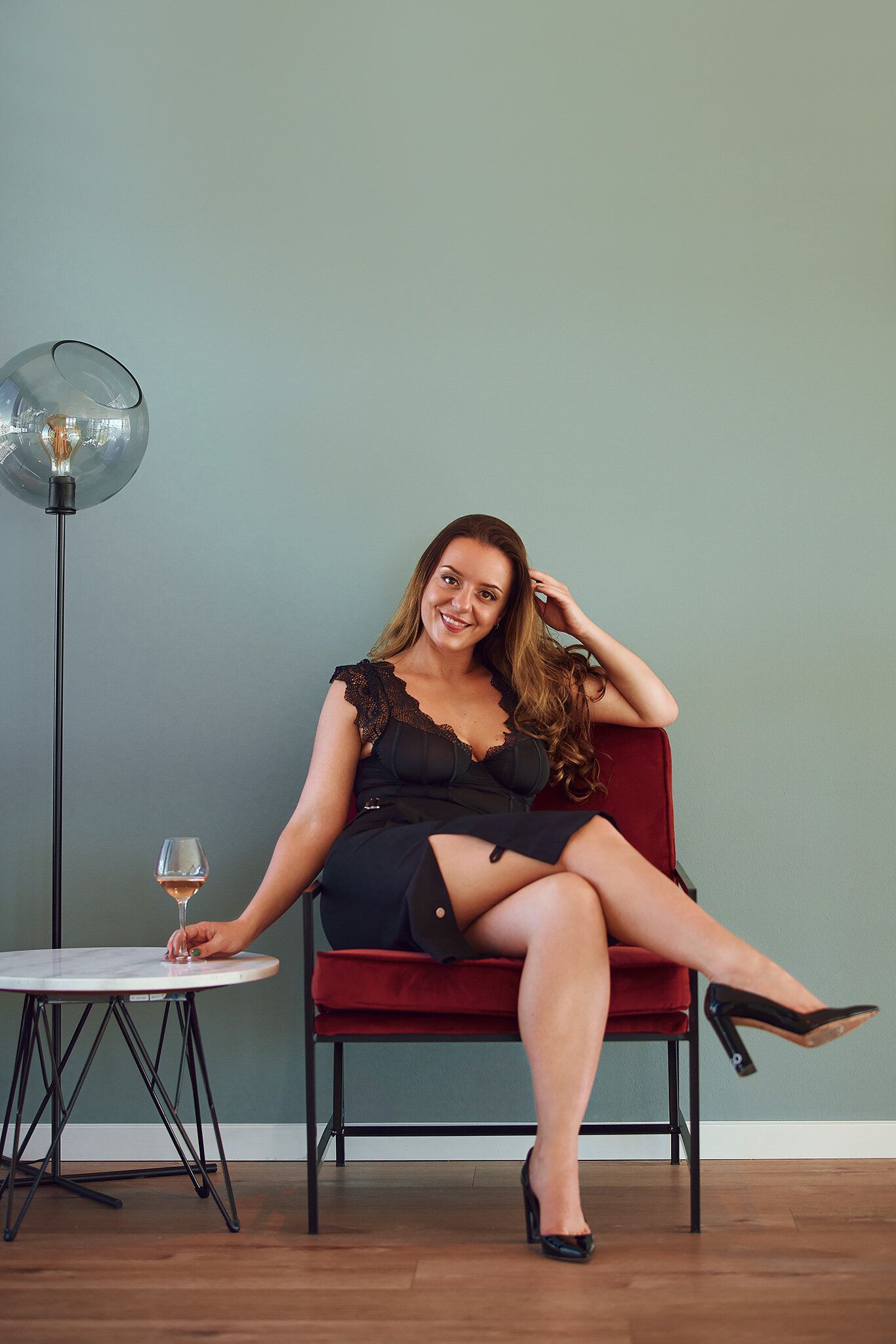 Woman in black dress sits in red chair, holding drink, smiles, room with lamp and table.