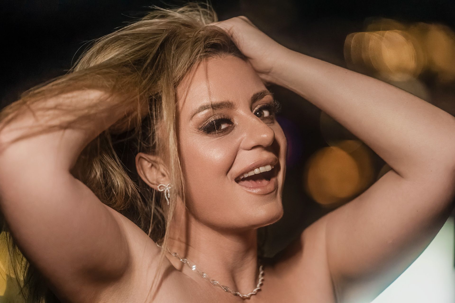Woman with light-colored hair, hands in hair, smiling with bright makeup and jewelry. Blurred background with lights.