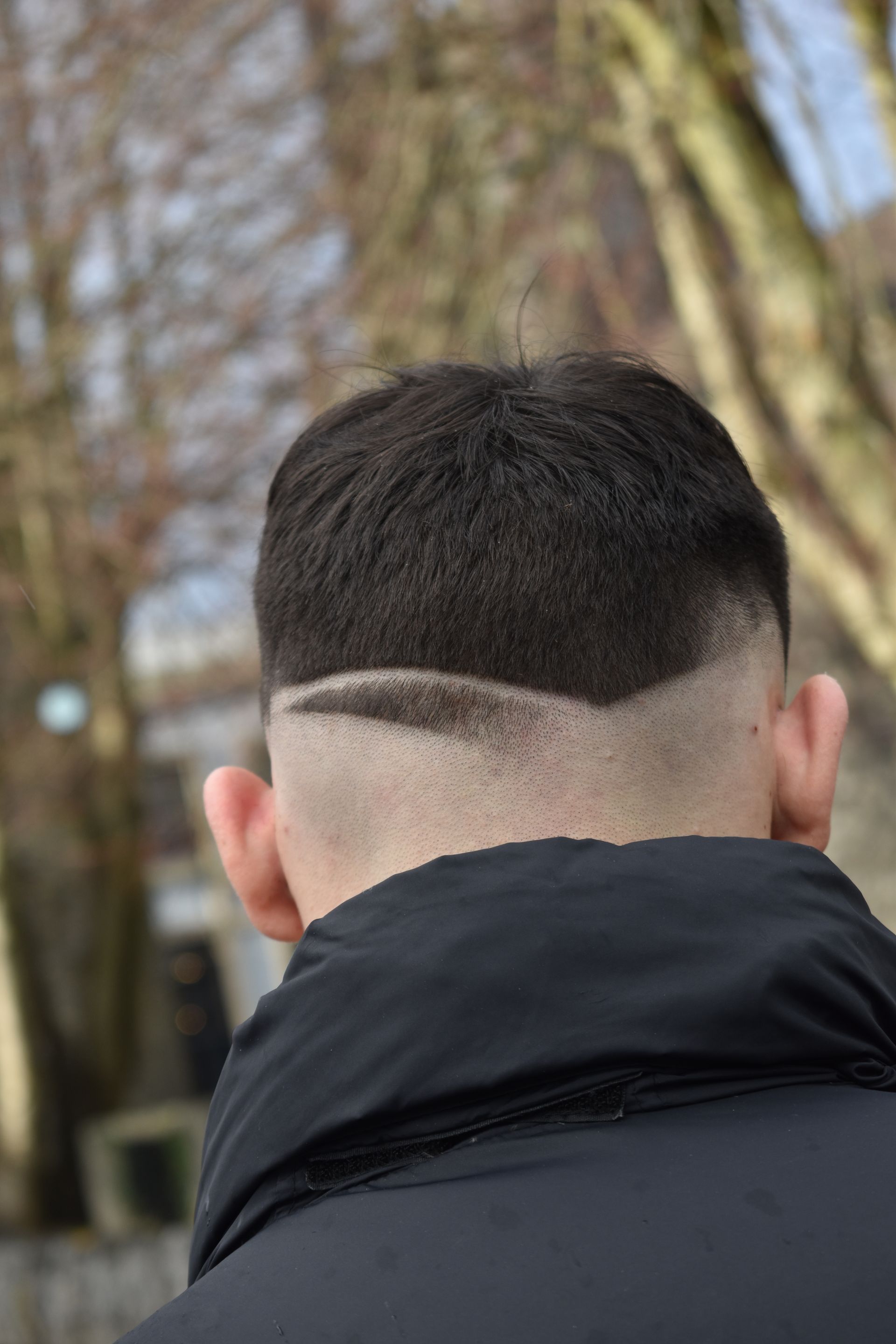 Person's dark hair with a faded undercut. A design is shaved into the nape. Blurred outdoor background.