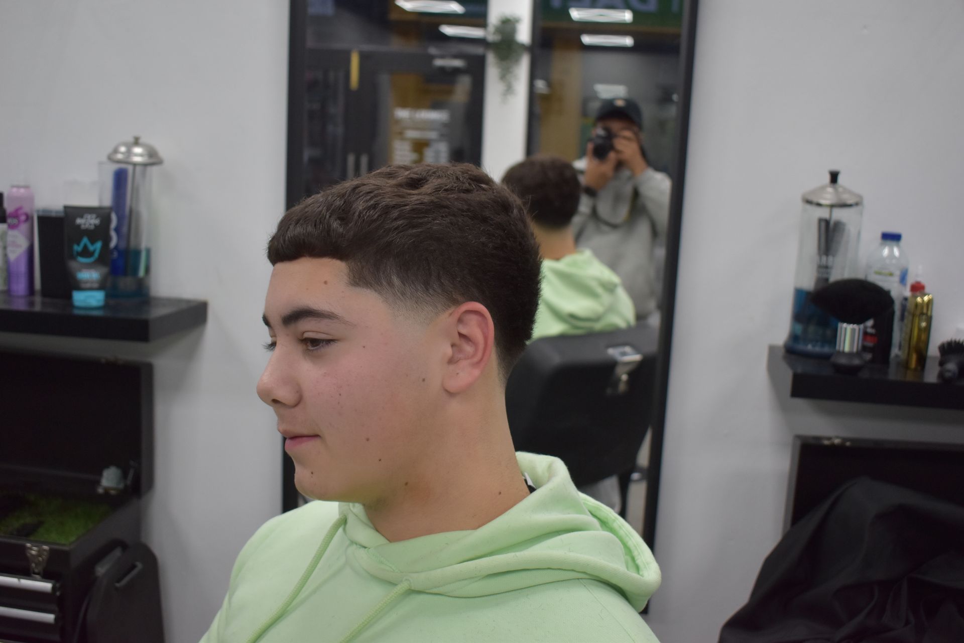 Person in a barber shop, profile view showing fresh haircut. Wearing a light green hoodie, with dark hair, and smiling slightly.