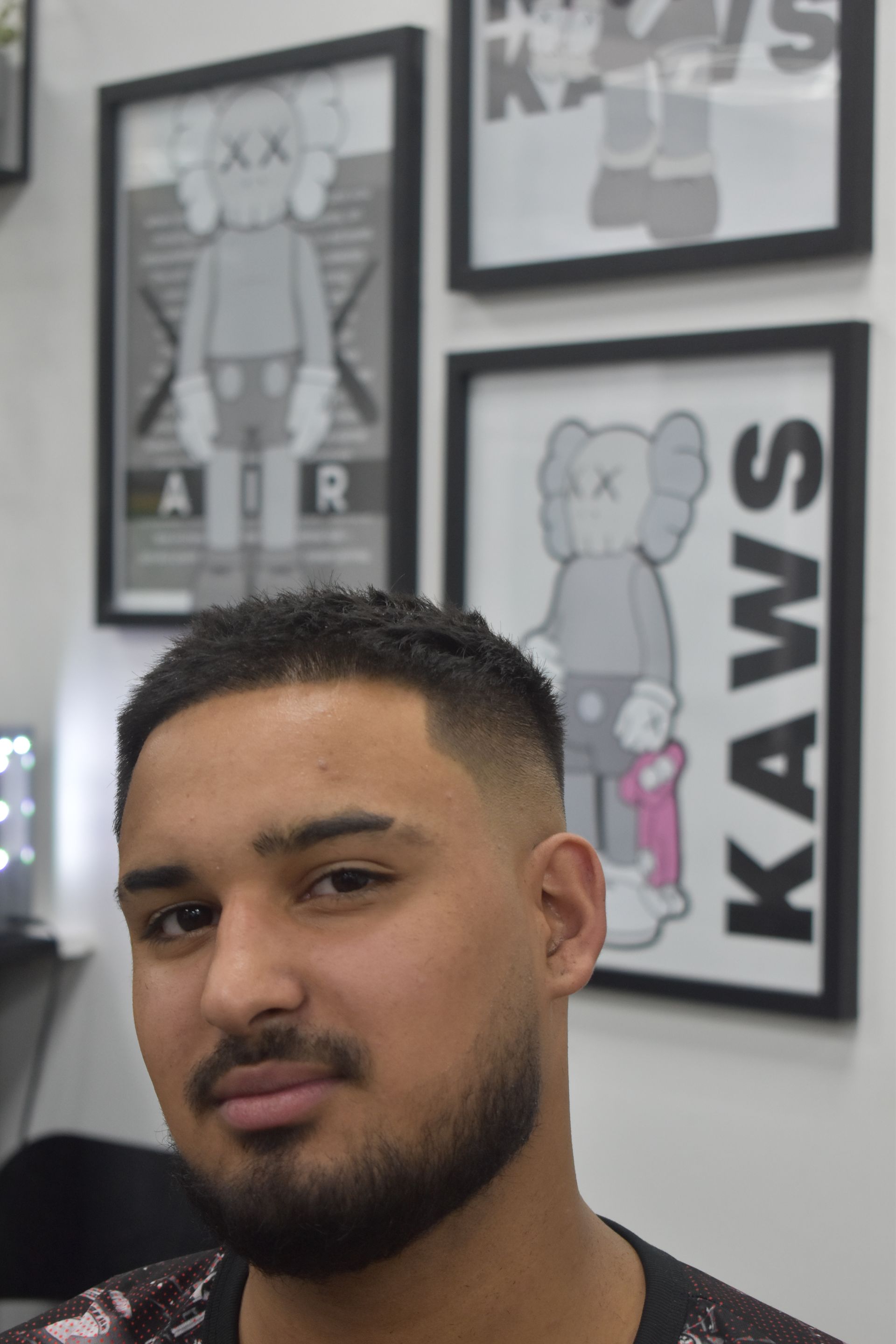 Man with faded haircut, small beard, smiling, posing in a room with KAWS art.