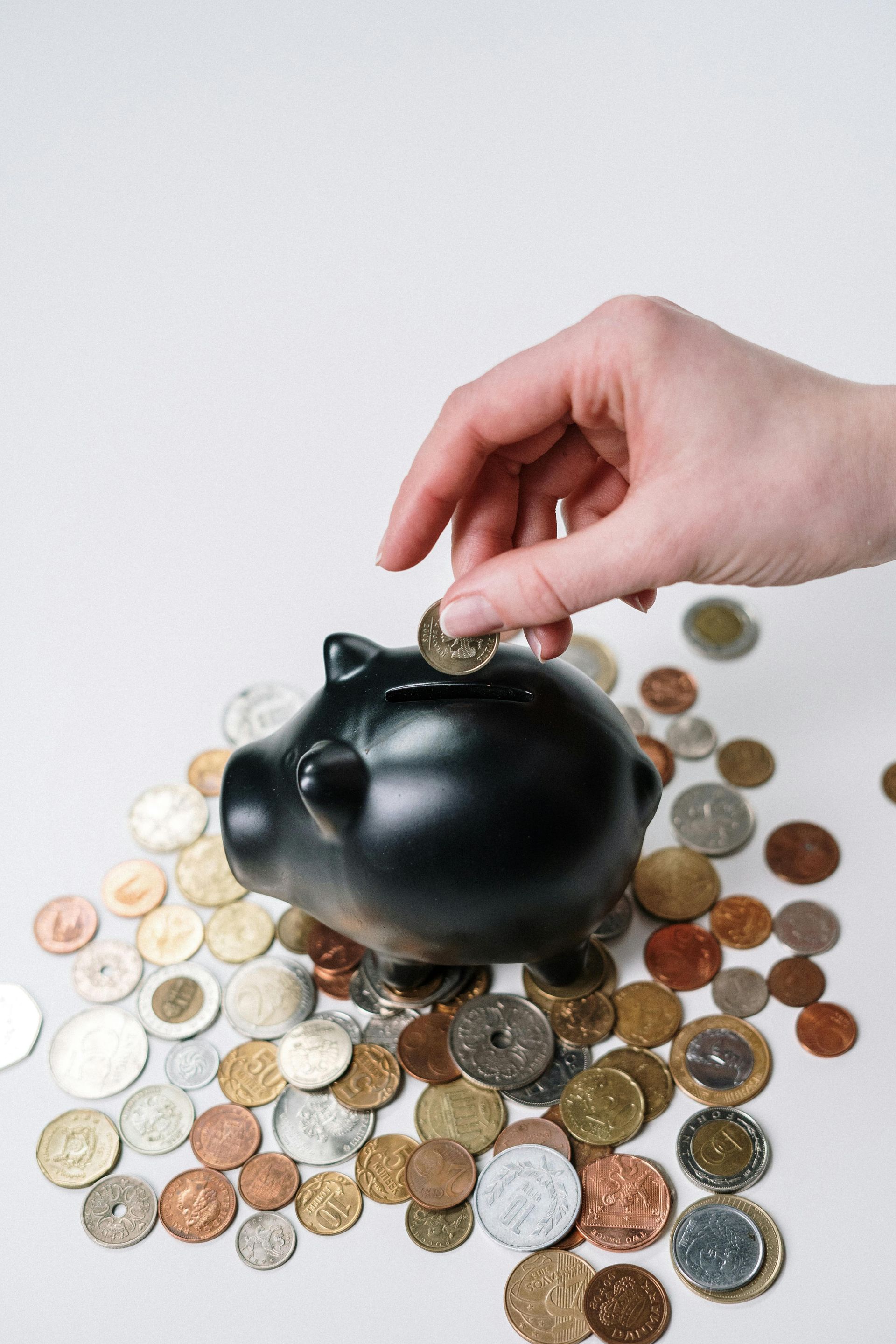 Hand placing a coin into a black piggy bank surrounded by scattered coins