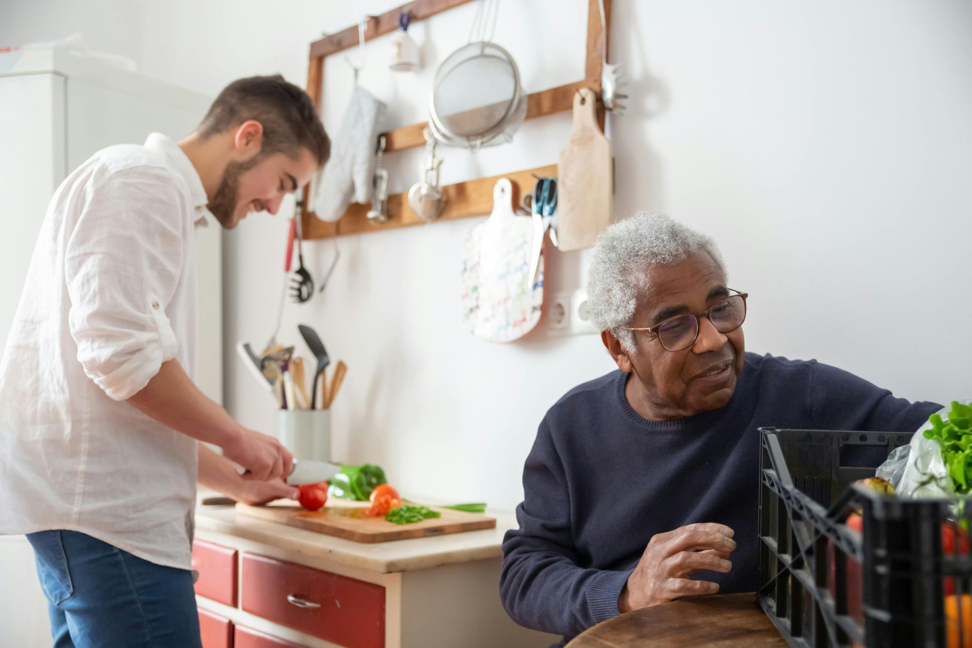 Two people preparing food in a bright kitchen, with one chopping vegetables and the other seated nearby.