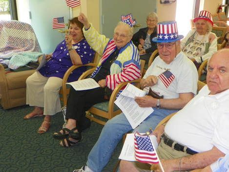 People seated in a room, wearing patriotic red, white, and blue outfits and holding small U.S. flags.