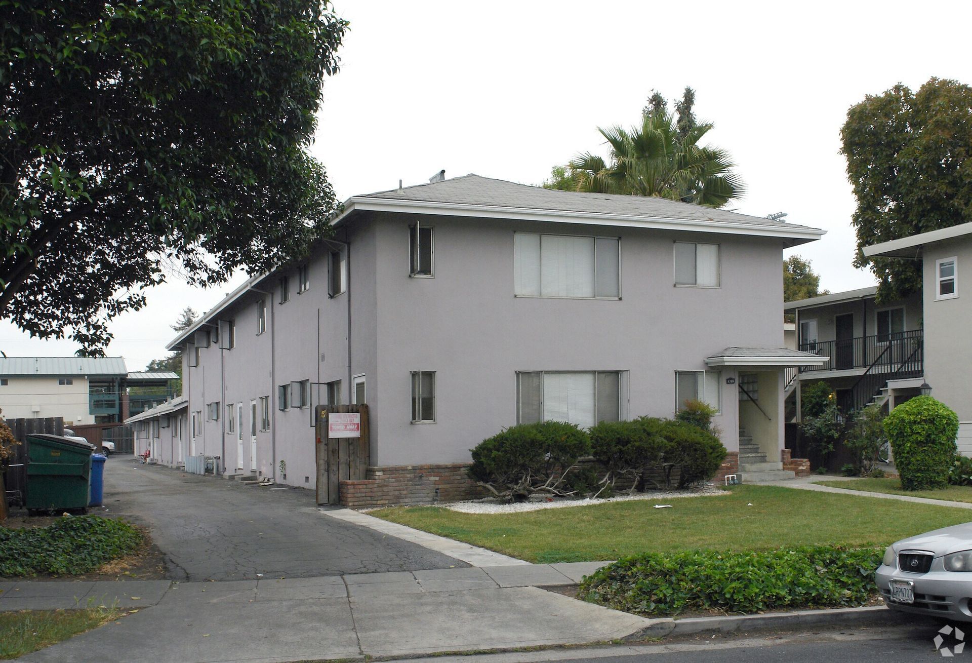 Gray two-story apartment building with a small lawn, driveway, and parked car on a cloudy day.