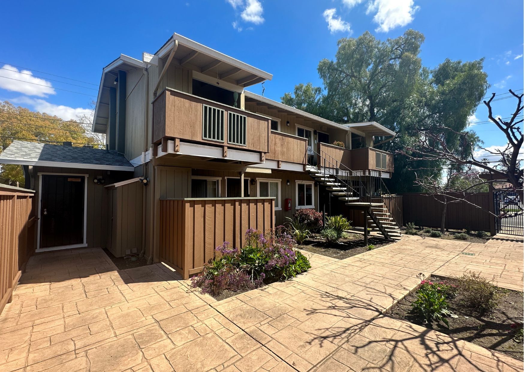 Two-story apartment building with brown balconies and stairs, surrounded by a brown fence and paved patio.