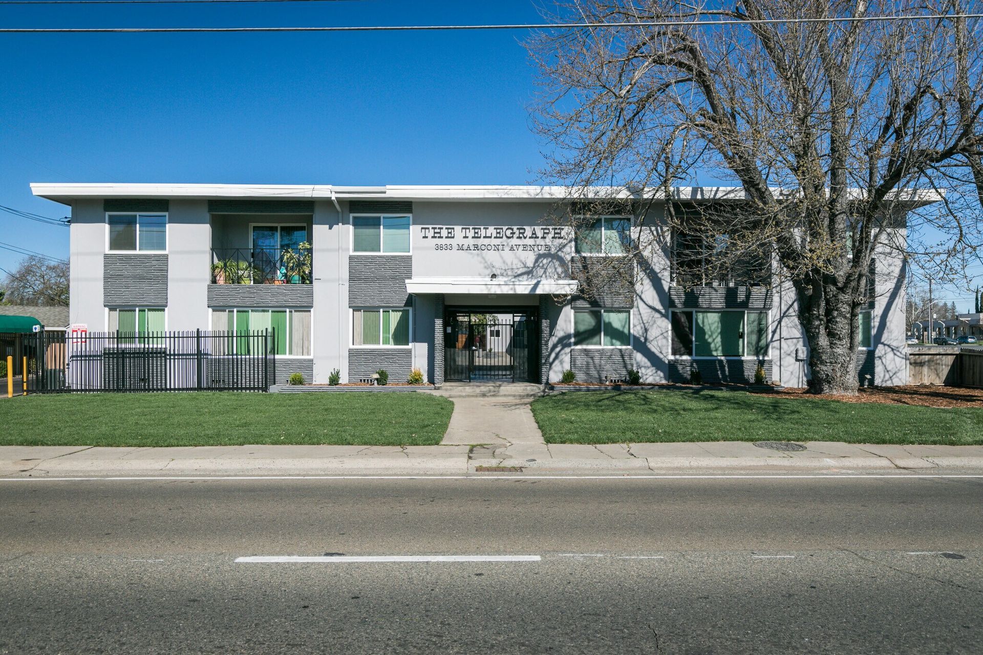 Two-story apartment building with a sidewalk, green lawn, and a tree on a sunny day.