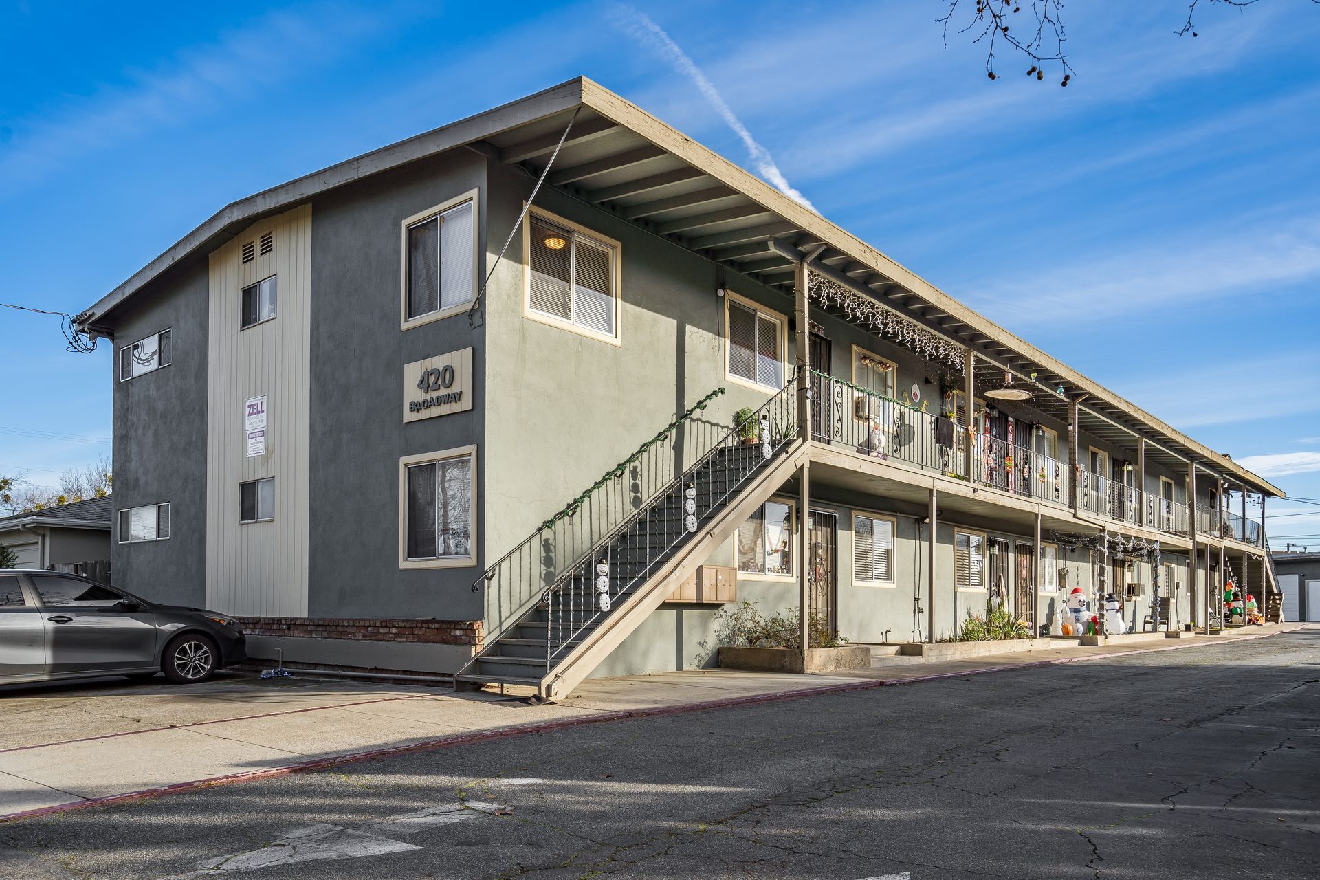 Two-story apartment building with gray siding, black staircases, and parked car on a sunny day.