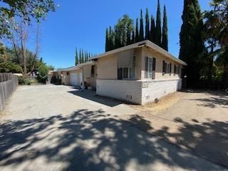 Tan and white one-story house with a long driveway under a blue sky, tall trees in the background.