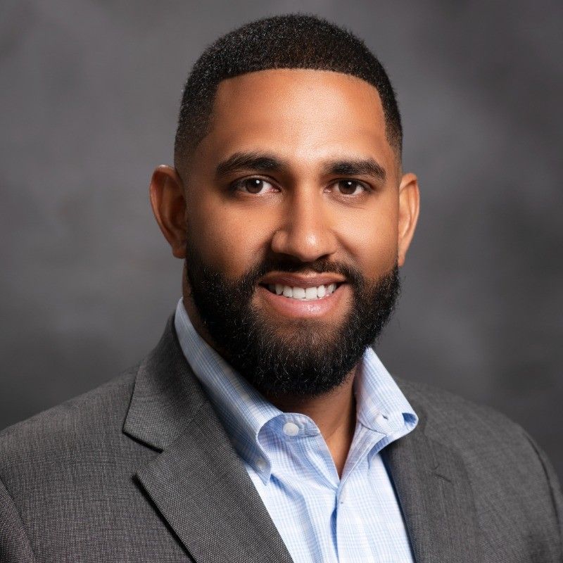 Man in gray suit and blue shirt smiles, headshot against a gray backdrop.