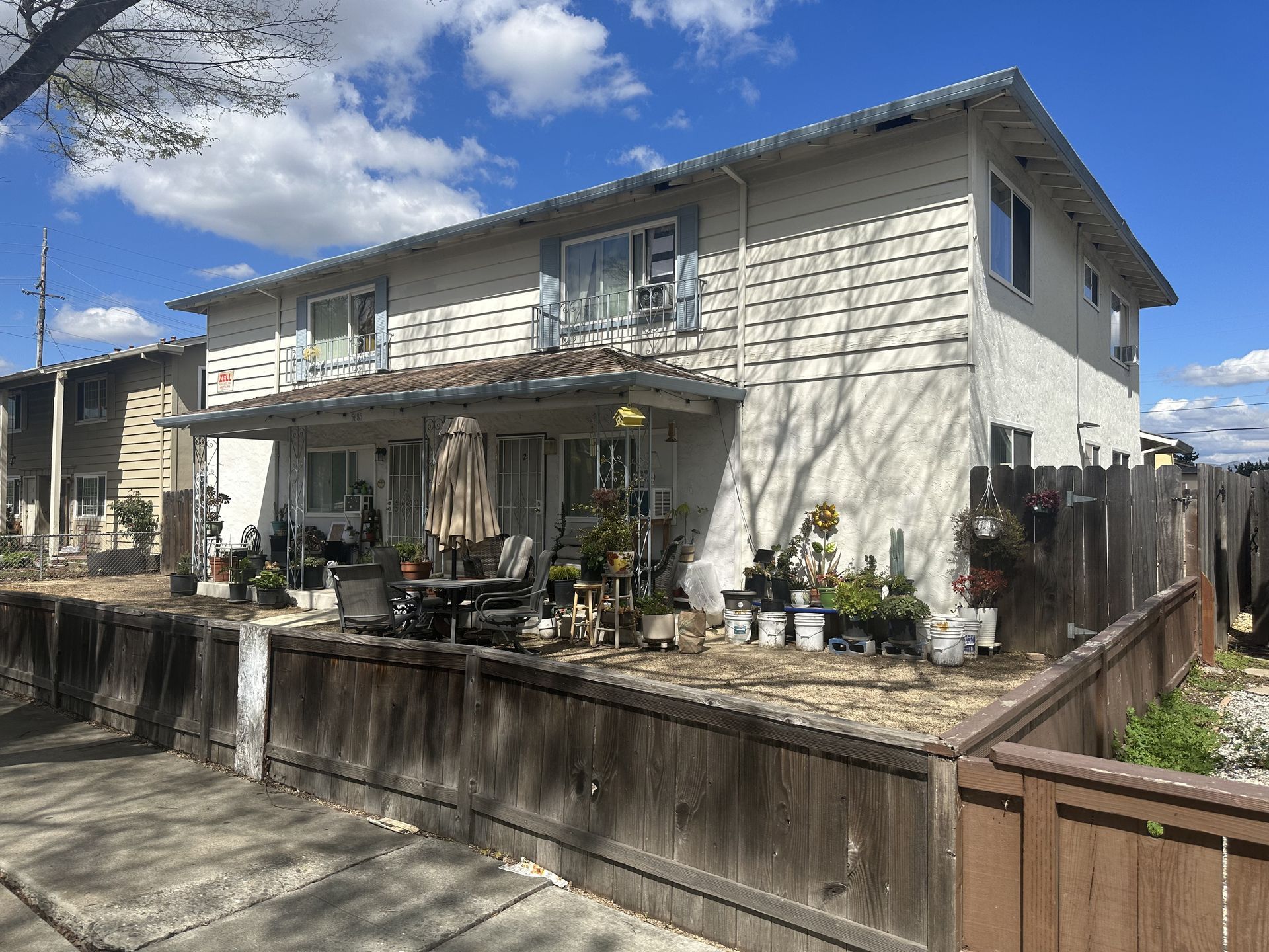 Two-story beige residential building with a wooden fence and various potted plants. Overcast sky.