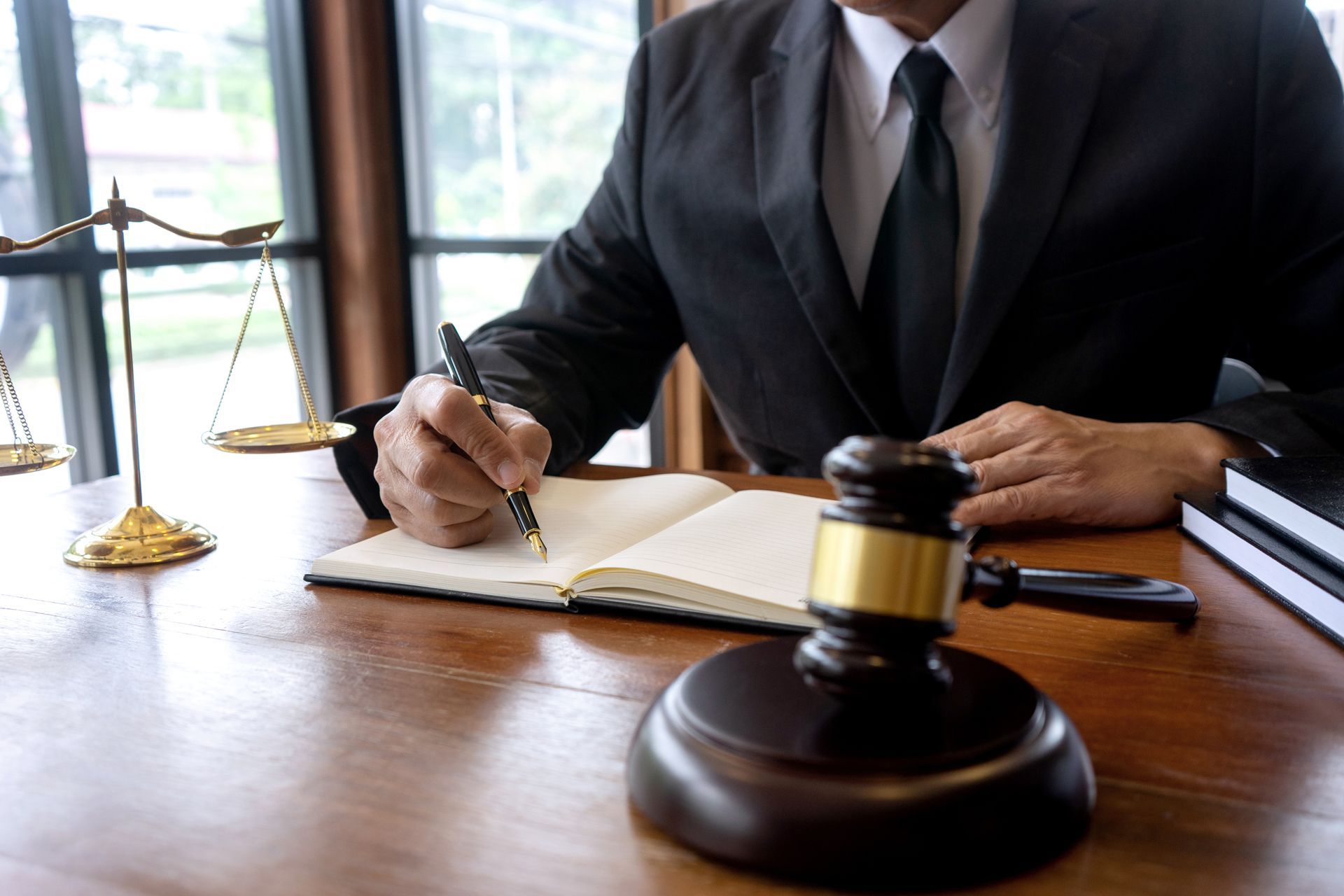 A lawyer in a suit writing in a notebook at a desk with a gavel, scales of justice, and books
