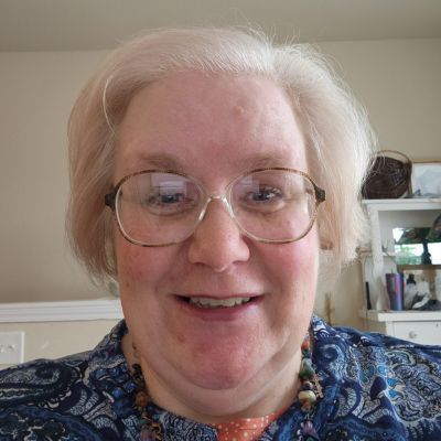 Woman with glasses smiles; wearing patterned blue shirt and beaded necklace; indoor setting.