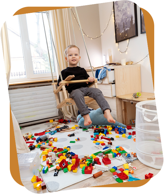 A child sitting on a wooden indoor swing in a room filled with colorful plastic building blocks scattered on the floor.