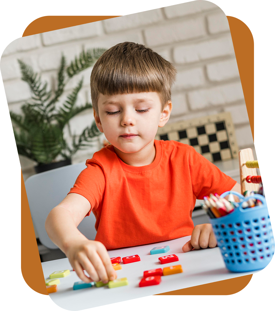 A child in an orange shirt sits at a desk, arranging colorful plastic letters on a white surface.