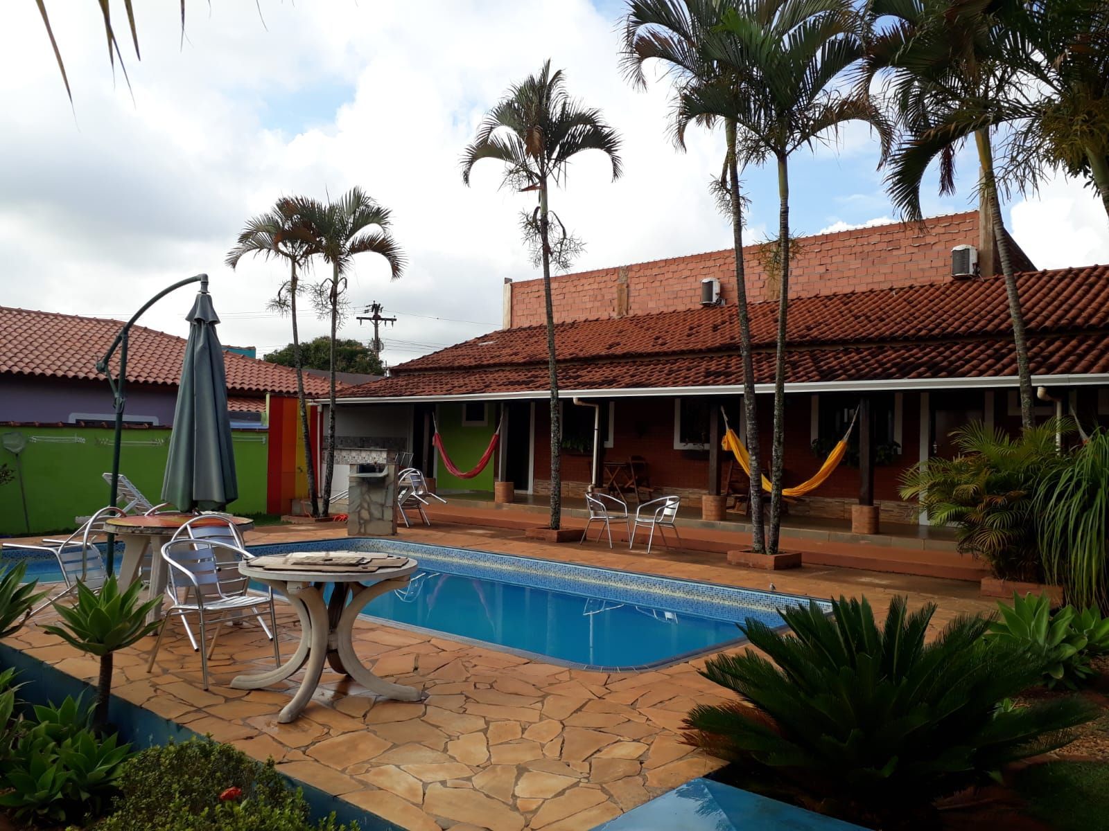Piscina de um resort ensolarado, com palmeiras, deck revestido de azulejos e cabanas de telhado vermelho refletidas na água