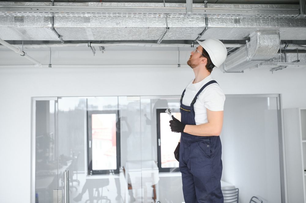 Man in overalls and hard hat inspecting ceiling ventilation in a bright room.
