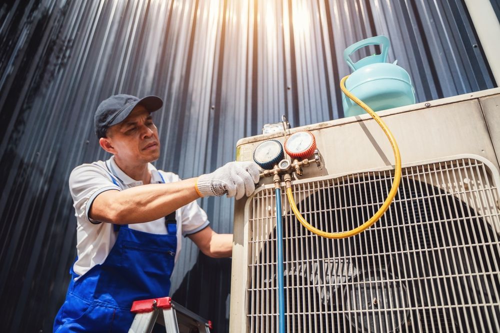 HVAC technician in overalls, attaching gauges to an AC unit, near a blue refrigerant tank.