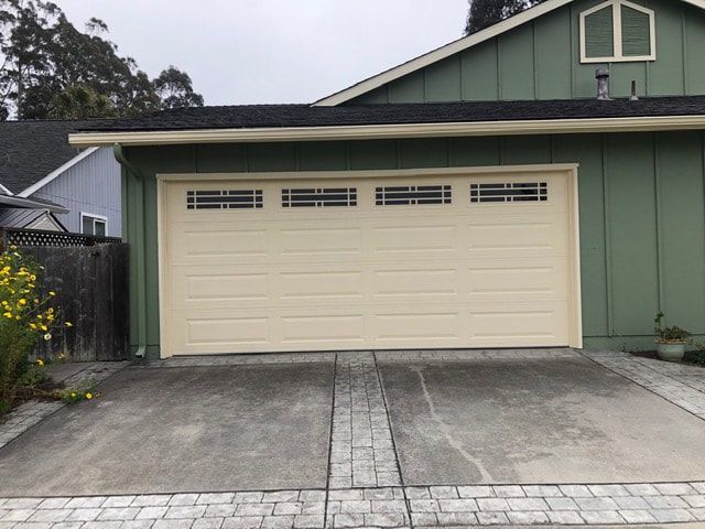 Beige double garage door on a green house with a concrete driveway and small side gate.