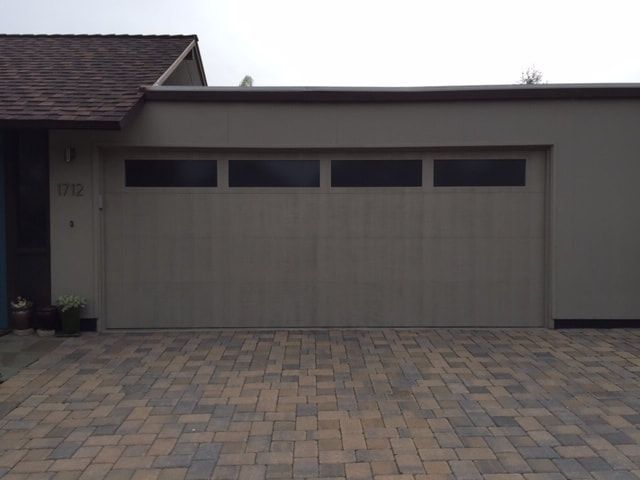 Gray garage door with three narrow windows, beside a house and paver driveway.