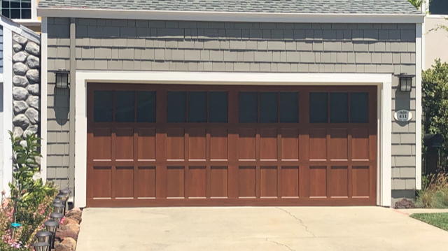 Brown double garage door with dark windows on a gray house exterior, above a concrete driveway