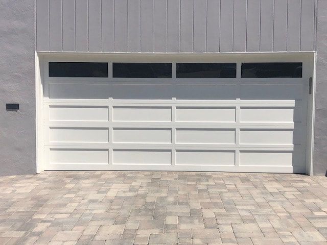 White garage door with four dark windows set in a gray wall above a stone-paved driveway