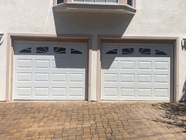 Two white garage doors on a beige house, with a brick driveway in front.