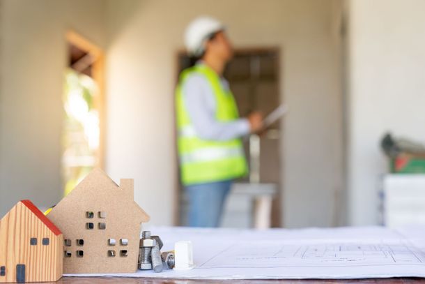 Small model houses sit on building plans in the foreground, with an inspector in a hard hat and vest blurred in the back.