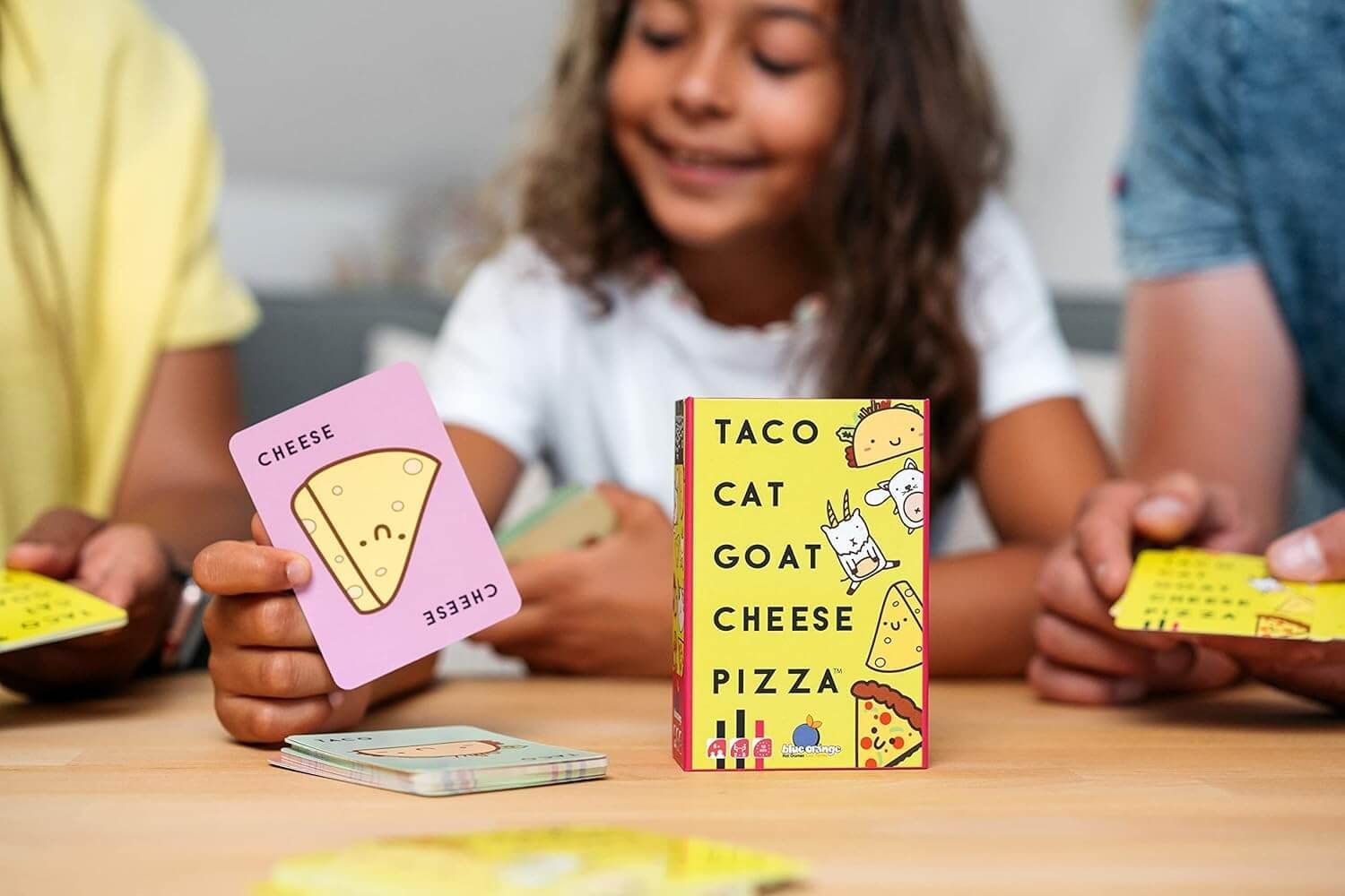 People playing Taco Cat Goat Cheese Pizza card game, showing cards and box on a wooden table.