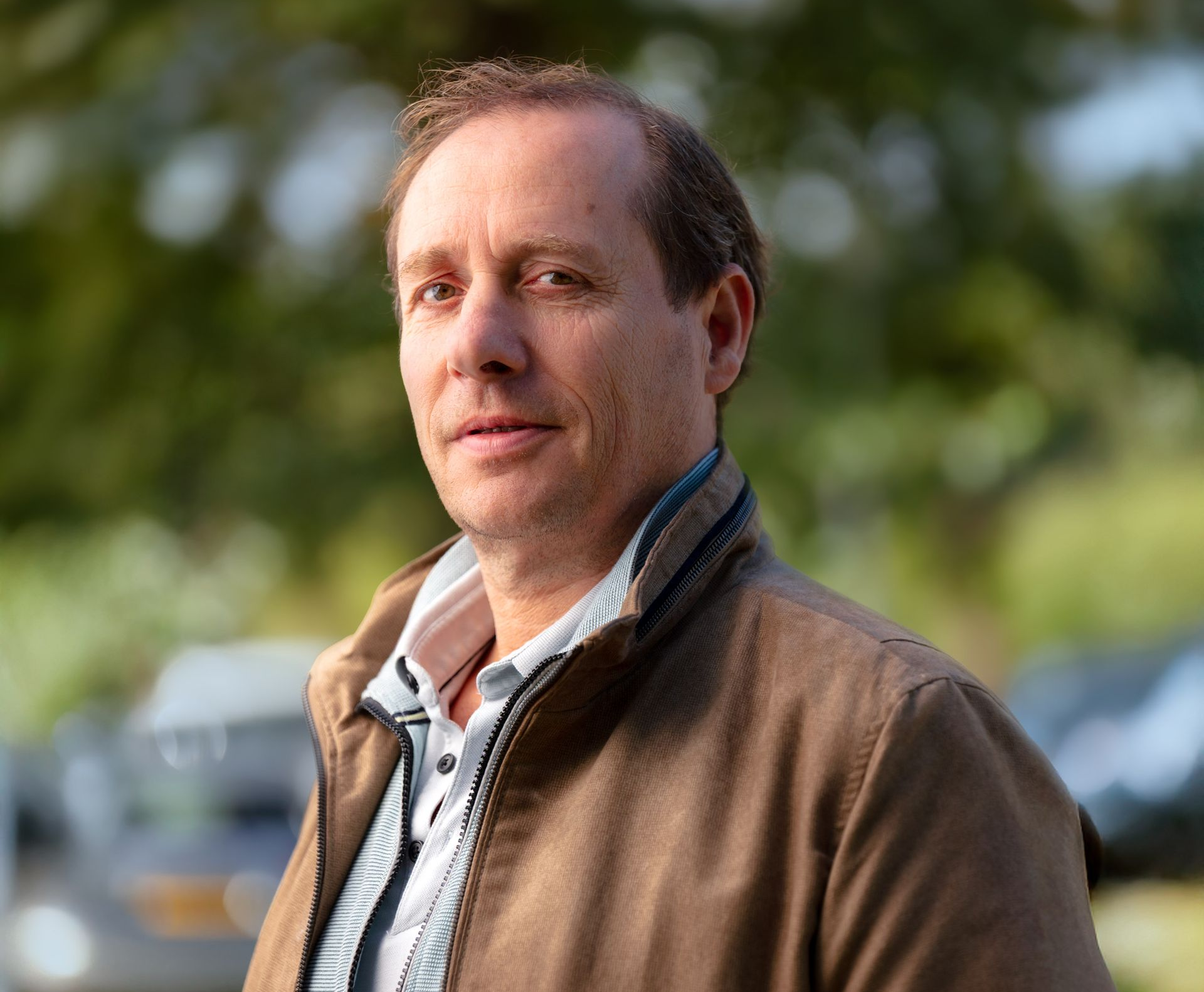 Portret van een man in een bruine jas, buiten, met een wazige groene achtergrond.