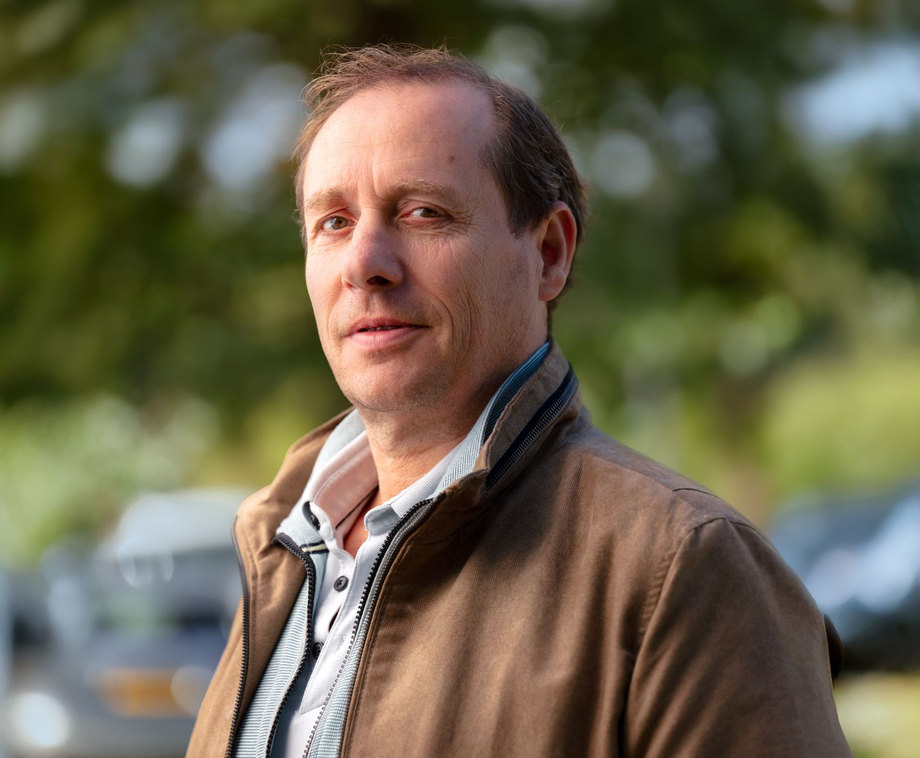 Portret van een man in een bruine jas, buiten, met wazig groen op de achtergrond.