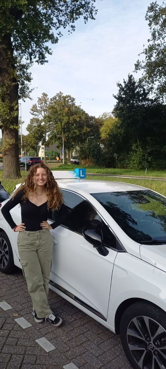 Vrouw staat naast een witte auto in een park, met bomen en gras op de achtergrond.