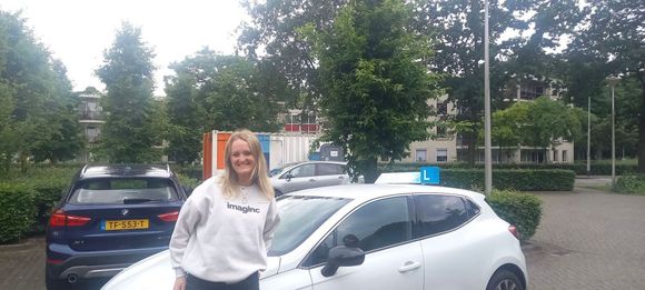 Vrouw staat naast een witte auto op een parkeerplaats met bomen en andere auto's op de achtergrond.