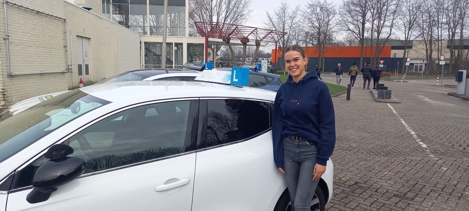 Een persoon staat naast een witte auto op een parkeerplaats en houdt een blauw bord op het dak vast.