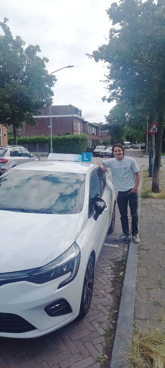 Een persoon staat naast een witte auto op een stoep onder bomen, met bakstenen gebouwen op de achtergrond.