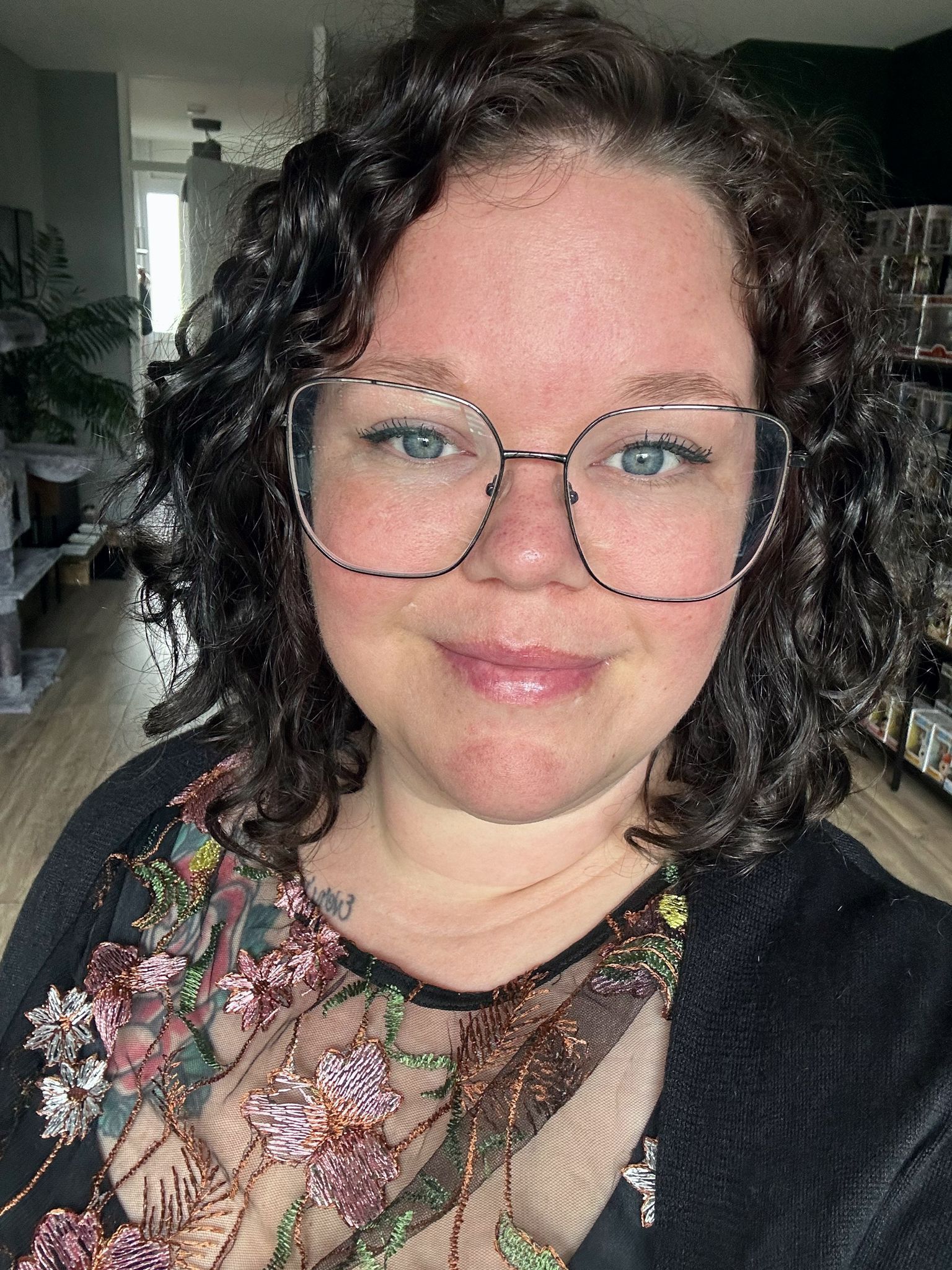 Een lachend persoon met een bril en krullend haar, gekleed in een top met bloemenprint, staat binnen.