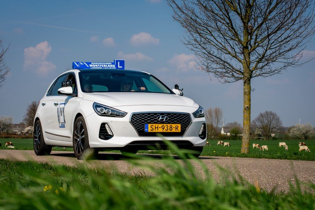 Een witte lesauto van een rijschool staat geparkeerd op een landweggetje, tussen gras en bomen, onder een blauwe hemel.