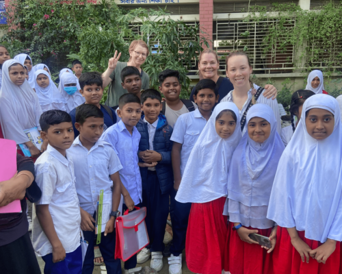 Group of students in white uniforms gathered outdoors with two adults, smiling and posing together